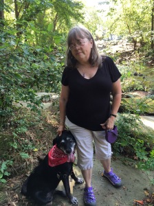 A woman with shoulder length hair wearing a black t-shirt, next to a black dog with a red bandana around its neck.