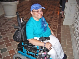 A person in a turquoise t-shirt and blue baseball cap and glasses sitting in a power chair